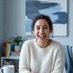 A diverse woman in her late 20s smiles warmly at the camera in a home office setting.