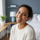 A happy, diverse woman in her late 20s to early 30s smiles warmly in a natural light portrait. The background is a blurred, c